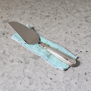 May include: A silver cake server with a serrated edge and a decorative handle featuring a shell motif at the base. The server rests on a light blue fabric pouch with the partial text TIFFANY visible in black lettering on the fabric surface.
