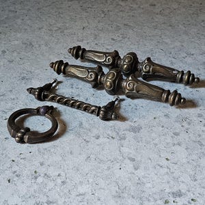 May include: A collection of antique bronze drawer pulls and a ring pull. The drawer pulls have ornate designs with spiral and scroll details. The ring pull is a simple, rounded design. All pieces have a vintage, aged appearance.
