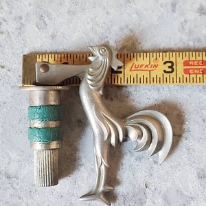 Puede incluir: Un abridor de botellas de plata con forma de gallo, con un mango verde y plateado. El abridor mide 7,6 cm de largo y se mide con una regla.