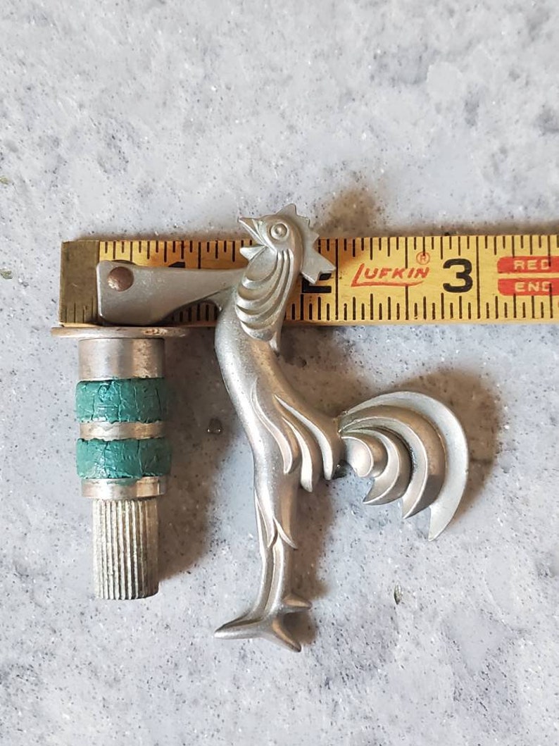Puede incluir: Un abridor de botellas de plata con forma de gallo, con un mango verde y plateado. El abridor mide 7,6 cm de largo y se mide con una regla.
