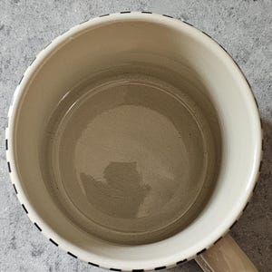 May include: Cream-colored ceramic mug with a rounded shape and a handle. The rim is decorated with a black dashed line. The interior of the mug is a light beige color.