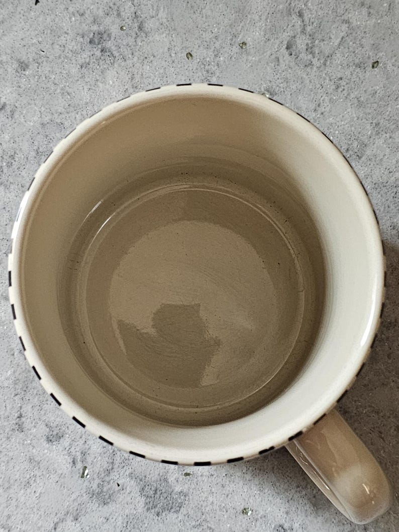 May include: Cream-colored ceramic mug with a rounded shape and a handle. The rim is decorated with a black dashed line. The interior of the mug is a light beige color.