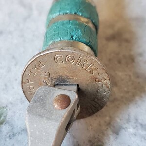 Puede incluir: Primer plano de un abridor de botellas plateado con un tap&oacute;n de corcho verde. El tap&oacute;n tiene las palabras "Cork-a-Pop" impresas.
