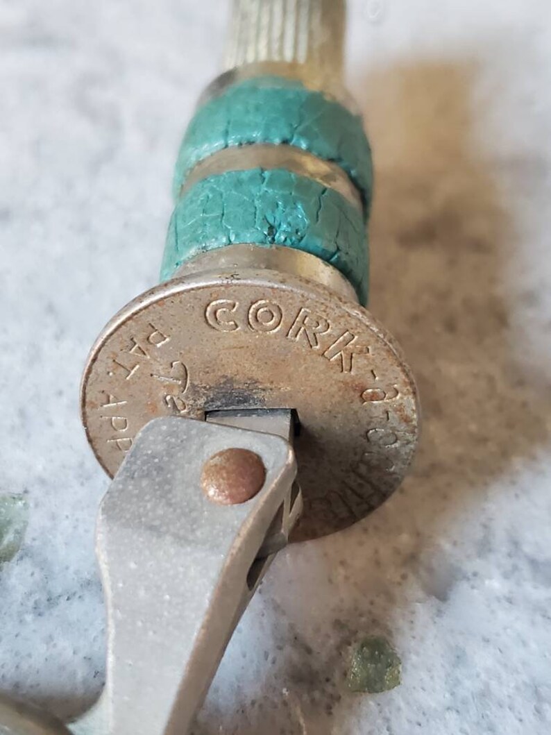 Puede incluir: Primer plano de un abridor de botellas plateado con un tap&oacute;n de corcho verde. El tap&oacute;n tiene las palabras "Cork-a-Pop" impresas.