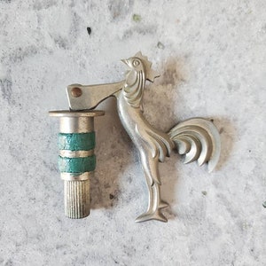 Puede incluir: Un abridor de botellas de plata con forma de gallo, con un mango verde y plateado. El gallo est&aacute; de pie sobre una superficie plana.
