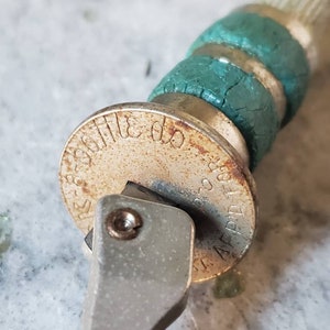 Puede incluir: Primer plano de un abridor de botellas plateado con una empu&ntilde;adura de goma verde. El abridor de botellas tiene una base circular con el texto "APPLIED FOR" y "220 300" impreso en &eacute;l.