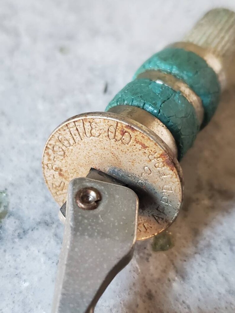 Puede incluir: Primer plano de un abridor de botellas plateado con una empu&ntilde;adura de goma verde. El abridor de botellas tiene una base circular con el texto "APPLIED FOR" y "220 300" impreso en &eacute;l.