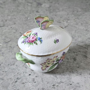 May include: A white porcelain sugar bowl with a lid and a green handle. The lid features a painted butterfly with pink and green wings. The bowl is decorated with a floral pattern in shades of pink, green, and yellow.
