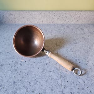 May include: A copper ladle with a wooden handle and a metal loop for hanging. The ladle's bowl is round and has a polished finish. The handle is light-colored wood.
