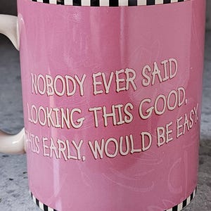 May include: Pink ceramic mug with the text "NOBODY EVER SAID LOOKING THIS GOOD THIS EARLY WOULD BE EASY" in white. The mug has a white handle and a black and white checkered border.