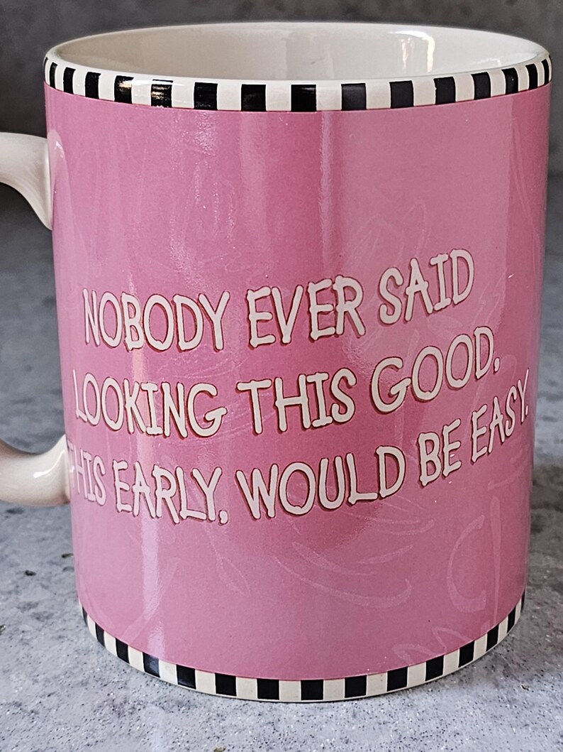 May include: Pink ceramic mug with the text "NOBODY EVER SAID LOOKING THIS GOOD THIS EARLY WOULD BE EASY" in white. The mug has a white handle and a black and white checkered border.