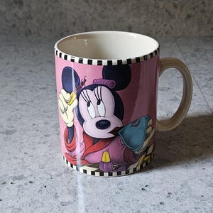 May include: Pink ceramic mug featuring Minnie Mouse in a spa-themed design. The mug has a cream-colored interior and handle, with a black and white checkered border. The design includes Minnie Mouse in a robe, holding a beauty item.