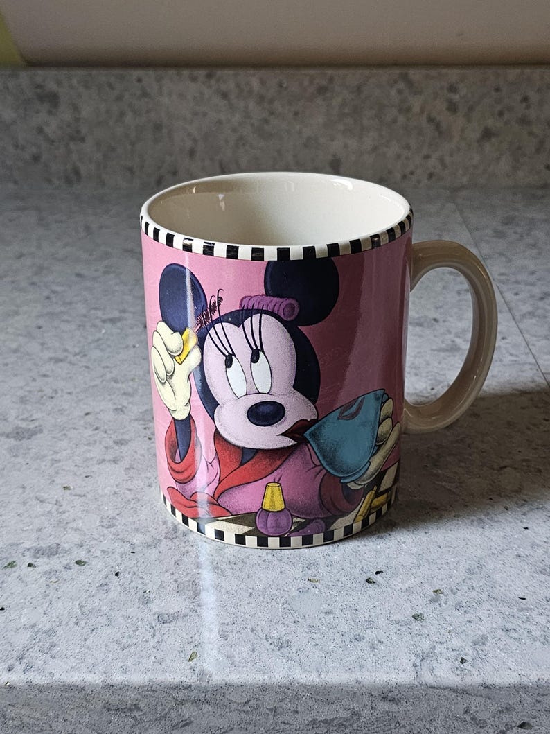 May include: Pink ceramic mug featuring Minnie Mouse in a spa-themed design. The mug has a cream-colored interior and handle, with a black and white checkered border. The design includes Minnie Mouse in a robe, holding a beauty item.