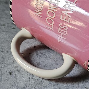 May include: Pink ceramic mug with the text "NOBODY LOOKING THIS EARLY". The mug has a white handle and a black and white striped rim. The Disney logo is visible on the side.