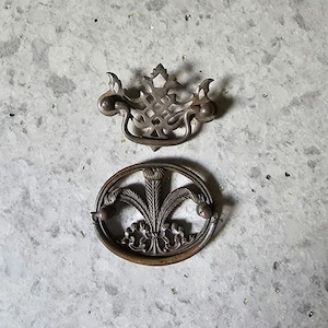 May include: Two antique brass drawer pulls. One is a simple oval shape with a decorative floral design. The other is a more ornate design with a square center and a decorative border.