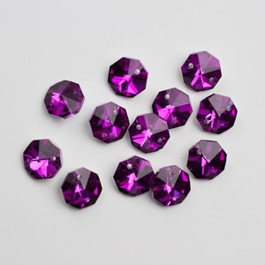 May include: A group of 14 purple crystal octagon beads with a faceted design. The beads are scattered on a white surface.
