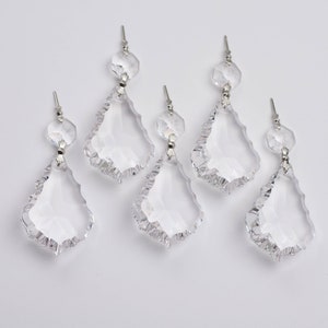 May include: Six clear crystal chandelier pendants with a teardrop shape and faceted edges. Each pendant is connected to a smaller, round crystal bead with a silver-colored metal pin. The pendants are arranged against a white background.