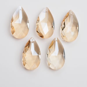 May include: Five teardrop-shaped crystal pendants with a faceted design. The crystals are a warm amber color and have a hole at the top for hanging. The pendants are arranged on a white background.