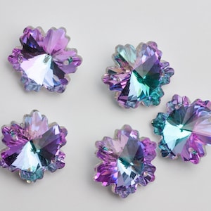 May include: Five iridescent, flower-shaped crystal embellishments. The crystals have a multi-faceted design with a color gradient of purple, blue, and green. They are arranged on a white background, showcasing their sparkling appearance.