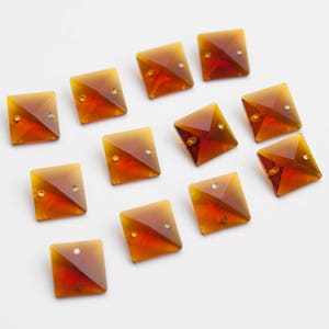 May include: A collection of twelve amber-colored square glass crystals with a pyramid shape. The crystals have a beveled edge and a small hole at the top.