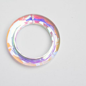 May include: Iridescent acrylic ring with a clear, faceted design. The ring reflects a spectrum of colors, including pink, orange, and purple. The ring is circular and has a hole in the center.