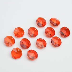 May include: A collection of eleven faceted, orange-red crystal beads. Each bead has multiple flat surfaces and two holes for stringing. The beads are scattered on a white surface, creating a visually appealing arrangement for jewelry making or crafting.
