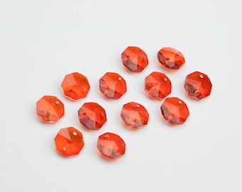 May include: A collection of eleven faceted, orange-red crystal beads. Each bead has multiple flat surfaces and two holes for stringing. The beads are scattered on a white surface, creating a visually appealing arrangement for jewelry making or crafting.