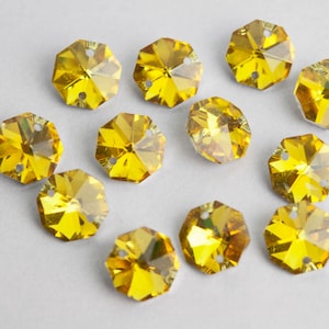 May include: A collection of 12 yellow faceted glass rhinestones with a hole in the center. The rhinestones are arranged in a heart shape on a white background.