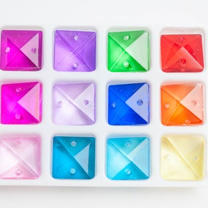 May include: A set of 10 square-shaped, faceted glass rhinestones in a variety of colors, including pink, purple, green, red, blue, orange, and yellow. The rhinestones are arranged in a grid pattern on a white background.
