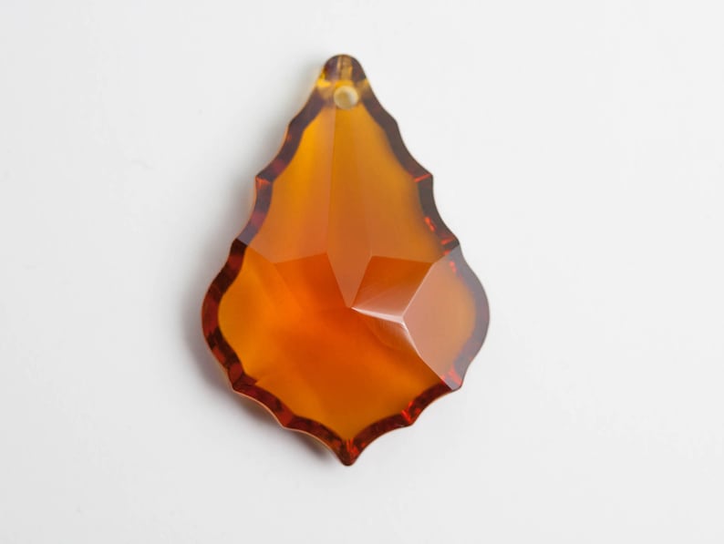 May include: A faceted, amber-colored crystal pendant with a teardrop shape. The pendant has a scalloped edge and a hole at the top for hanging. The crystal reflects light, showcasing its multiple facets.