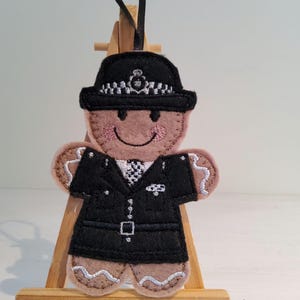 May include: A felt gingerbread man Christmas ornament in the shape of a police officer. The ornament features a black police hat and uniform, with a smiling face and pink cheeks. It is displayed on a small wooden easel and has a black ribbon.