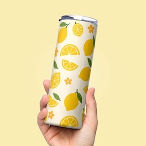 Könnte beinhalten: Ein weißer Becher mit einem Zitronen-Druck. Der Becher hat einen silbernen Deckel und wird in einer Hand gehalten.