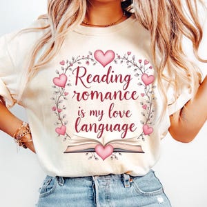 Può includere: T-shirt color crema con la scritta "Reading romance is my love language" in un design floreale a forma di cuore. Il design include cuori rosa e un'illustrazione di un libro. La maglietta è in materiale morbido.