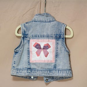 May include: A light blue denim vest with a decorative patch on the back. The patch features a pink and purple bow design within a white square, framed by a pink border. The vest is hanging on a beige hanger.