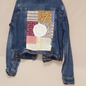 Puede incluir: Chaqueta vaquera azul con un diseño de patchwork en la espalda. El patchwork incluye varias telas en rojo, blanco, amarillo y azul, con un tapete blanco en el centro. La chaqueta está colgada en una percha.