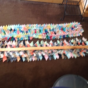 May include: Two colorful rag rugs made from strips of fabric, laid out on a brown carpet. The rugs are both about 1.5 feet wide and 4 feet long. The top rug is made from a variety of bright colors, including blue, green, yellow, and pink. The bottom rug is made from a variety of muted colors, including gray, brown, and beige. A wooden dowel rod is placed between the two rugs.