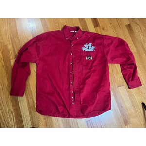Puede incluir: Una camisa abotonada de manga larga en un tono rojo vibrante. La camisa presenta un diseño bordado en el bolsillo del pecho, con una imagen de dibujos animados. La camisa tiene un cuello clásico y puños abotonados. La camisa está hecha de un material suave, probablemente algodón.