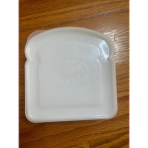 May include: A white plastic sandwich container shaped like a slice of bread. The container has a lid and the word "wonder" is printed on the front. The container is sitting on a wooden surface.