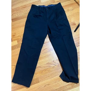 May include: Navy blue pleated trousers. The trousers are made of a durable fabric and have a button and zip fastening. The pants are displayed on a wooden floor.