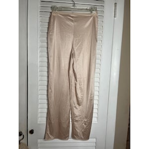 May include: A pair of beige, wide-leg trousers hanging on a hanger. The trousers are made of a shiny, silky material. The trousers are hanging against a white door.