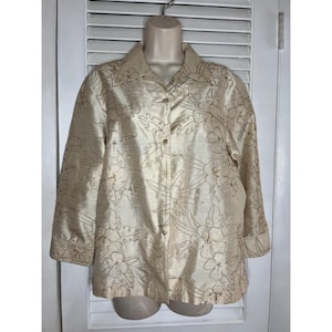 Può includere: Una camicetta color crema con bottoni e un motivo ricamato floreale. La camicia a maniche lunghe ha un colletto ed è esposta su un manichino. Il ricamo è in una tonalità crema più scura.