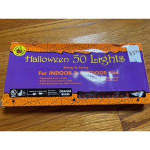 May include: A rectangular box of 50 orange Halloween lights for indoor and outdoor use. The purple packaging features a black spiderweb border, the text Halloween 50 Lights, and notes that the bulbs are replaceable and the string connects at both ends.