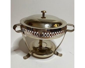 Può includere: Un chafing dish color argento con coperchio e una fascia traforata decorativa al centro. Il piatto ha due manici e poggia su un supporto con gambe curve. Un pomello color oro è sulla parte superiore del coperchio.