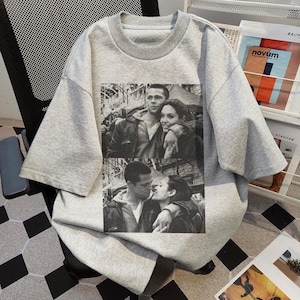 Könnte beinhalten: Ein hellgraues T-Shirt mit kurzen Ärmeln und einem Schwarz-Weiß-Bild eines Paares, das sich umarmt und küsst. Das Shirt hat einen Rundhalsausschnitt und eine lockere Passform. Das Bild ist eine Collage aus zwei separaten Fotos.