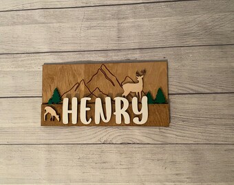 May include: Wooden sign with a mountain range, two deer, and the name "HENRY" in white letters.
