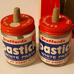 Puede incluir: Dos frascos de vidrio vintage de pasta blanca Pastic de Stafford para trabajos en papel. Cada frasco tiene una tapa roja con el texto "Since 1858" y un tapón de corcho. Las etiquetas son azules y blancas.
