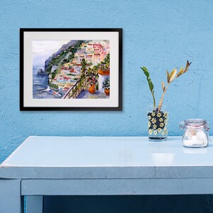 May include: A framed watercolor painting of a colorful Italian village on a cliffside overlooking the ocean. The painting is in a black frame and is hanging on a light blue wall.