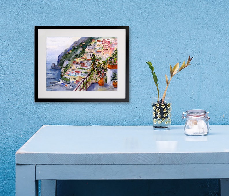 May include: A framed watercolor painting of a colorful Italian village on a cliffside overlooking the ocean. The painting is in a black frame and is hanging on a light blue wall.