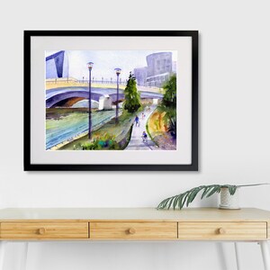 May include: Watercolor painting of a bridge over a river with a walking path and people walking and biking. The painting is framed in black.