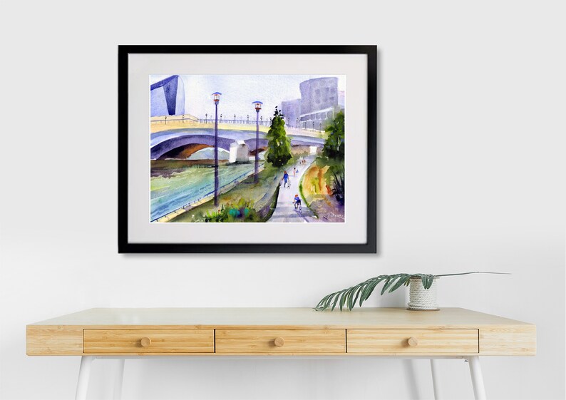 May include: Watercolor painting of a bridge over a river with a walking path and people walking and biking. The painting is framed in black.